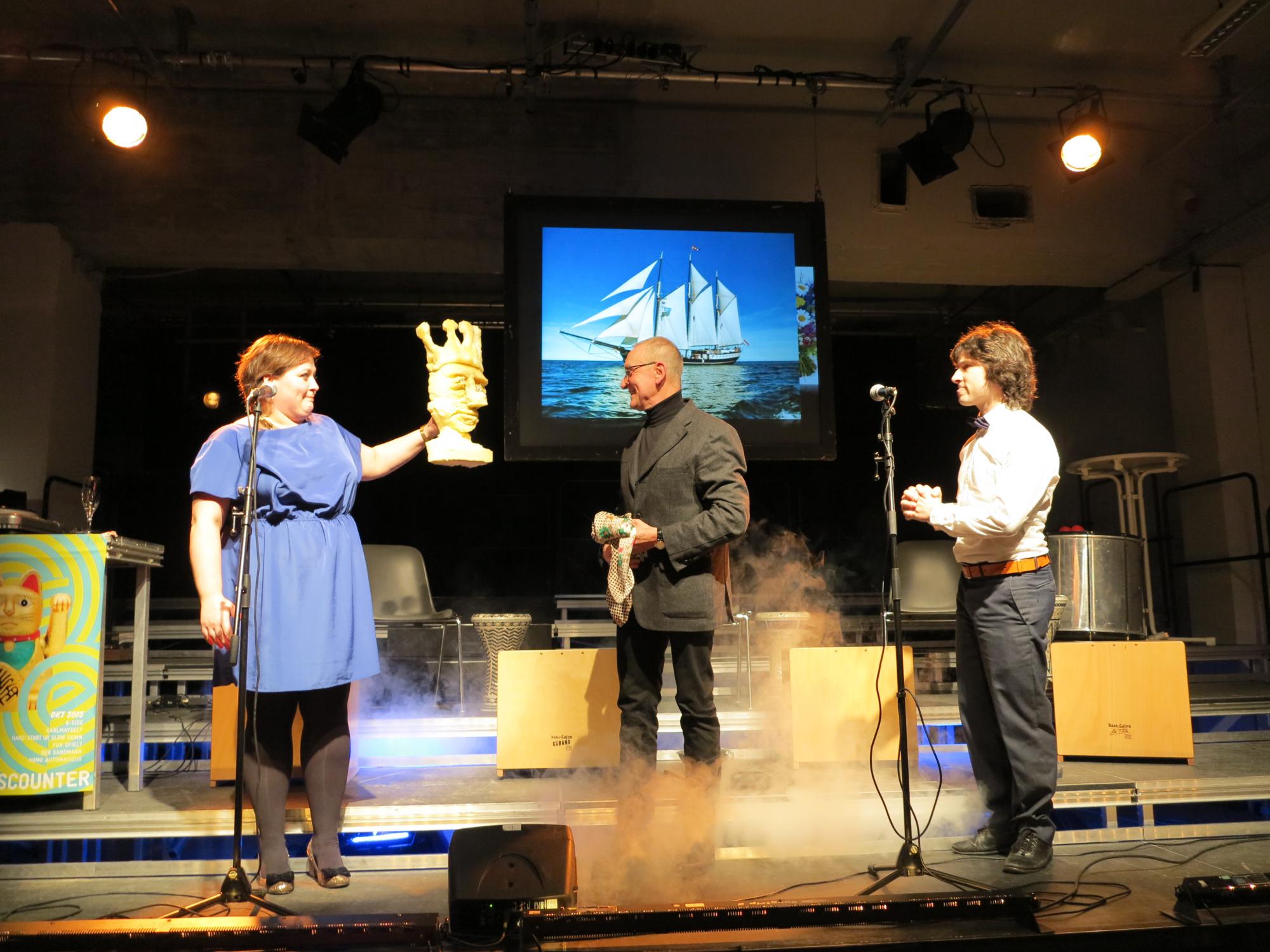 Die beiden Moderatoren der Feier Tina Pfurr und Martin Clausen überreichen Günter Jeschonnek eine Skulptur der Berliner Gruppe „Helmi“