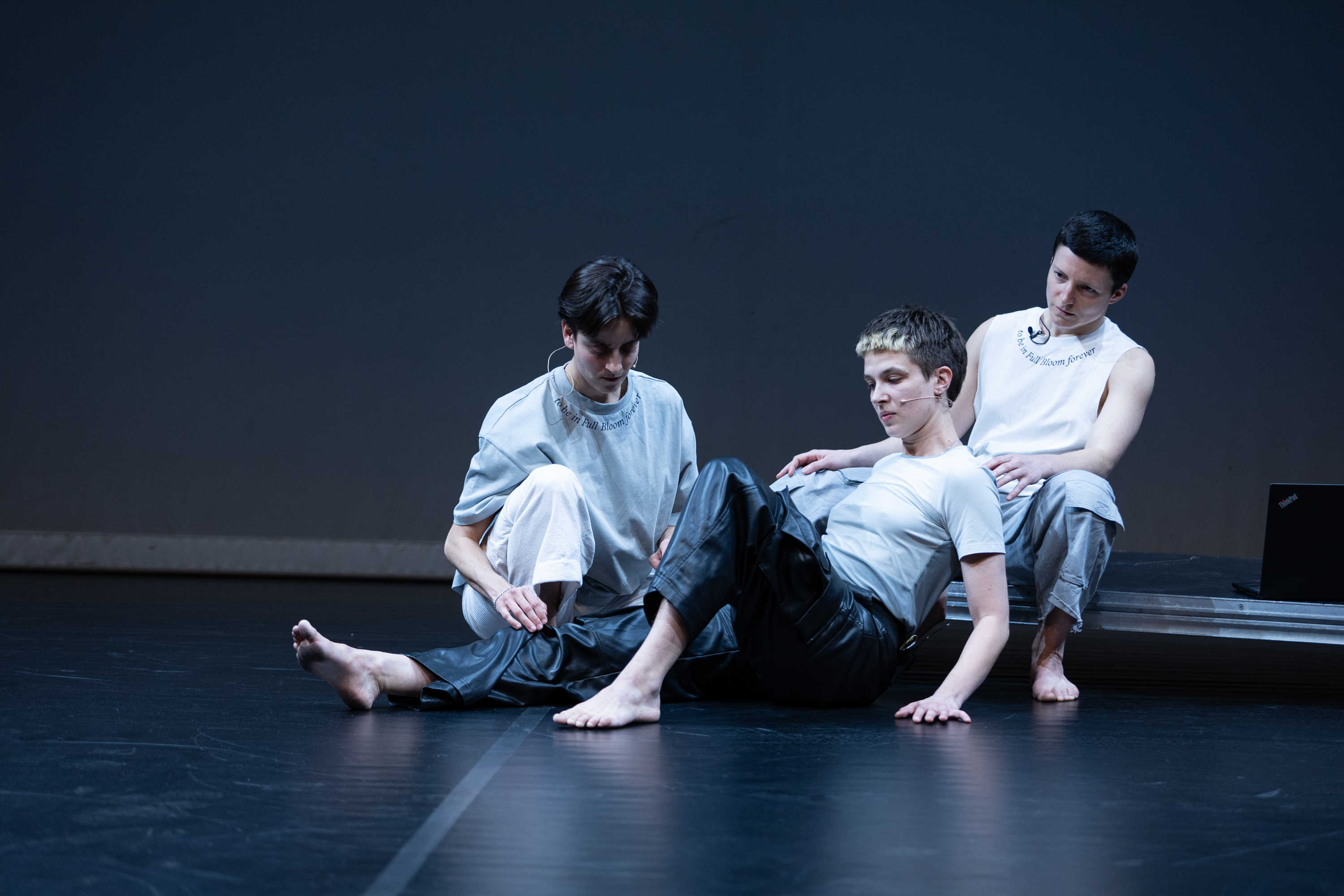 Drei Personen sitzen nebeneinander auf dem Boden. Sie halten eine ruhige, gemeinsame Pose, als wären sie Teil eines inszenierten Standbilds. Die Umgebung und ihre Kleidung erscheinen überwiegend in Grau-, Blau-, Weiß- und Schwarztönen.