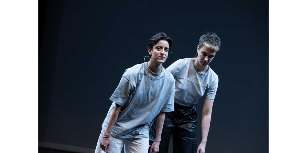 Zwei Personen stehen dicht nebeneinander, die Arme hängen nah am Körper. Sichtbar sind sie von den Oberschenkeln bis zum Kopf. Beide tragen T-Shirts und sportliche Hosen. Ihre Körper neigen sich leicht schräg nach rechts oben. Blau-, Grau-, Weiß- und Schwarztöne dominieren.