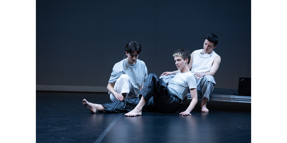 Drei Personen sitzen nebeneinander auf dem Boden. Sie halten eine ruhige, gemeinsame Pose, als wären sie Teil eines inszenierten Standbilds. Die Umgebung und ihre Kleidung erscheinen überwiegend in Grau-, Blau-, Weiß- und Schwarztönen.