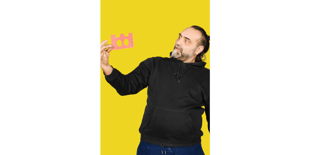 Ein Mann steht vor einer gelben Wand. Er hat dunkles Haar und einen ergrauenden Bart. Er trägt einen schwarzen Pullover und schaut erstaunt eine ausgeschnittene rosa Form in seiner Hand an.