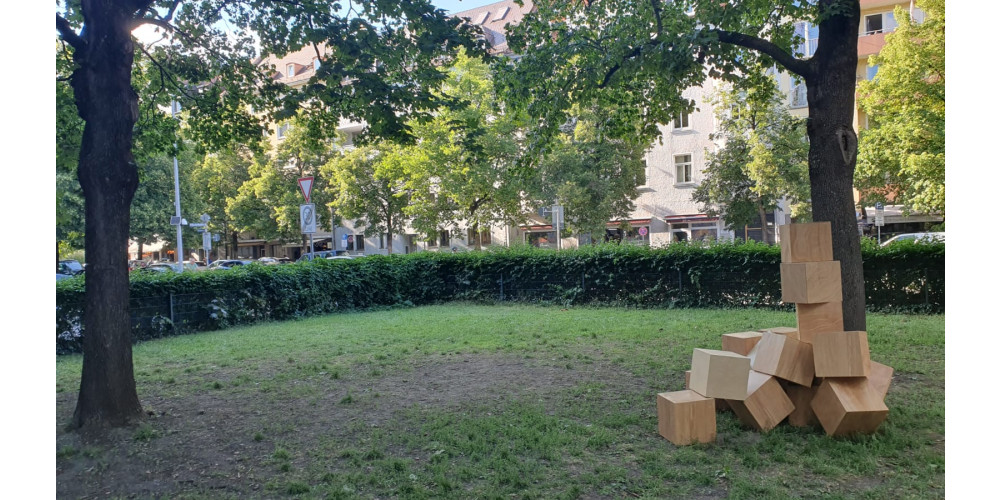 Foto: In einem Stadtpark sind Holzwürfel zu einer Art Skulptur aufgestapelt. Sie befindet sich auf der rechten Bildseite, die Mitte des Bildes zeigt eine ansonsten leere Stelle des Parks. Das Bild wird von zwei Bäumen gerahmt. Der Park ist durch eine Hecke abgetrennt. Im Hintergrund sind Straßenschilder und Häuser zu sehen.