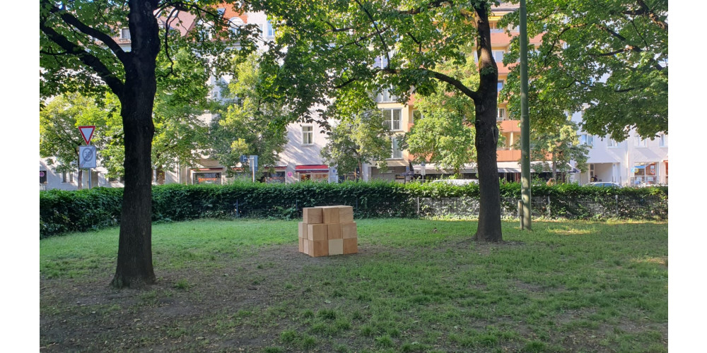 Foto: In einem Stadtpark sind zwischen zwei Bäumen Holzwürfel aufgestapelt. Der Park ist durch eine Hecke abgetrennt. Im Hintergrund sind Straßenschilder und Häuser zu sehen.