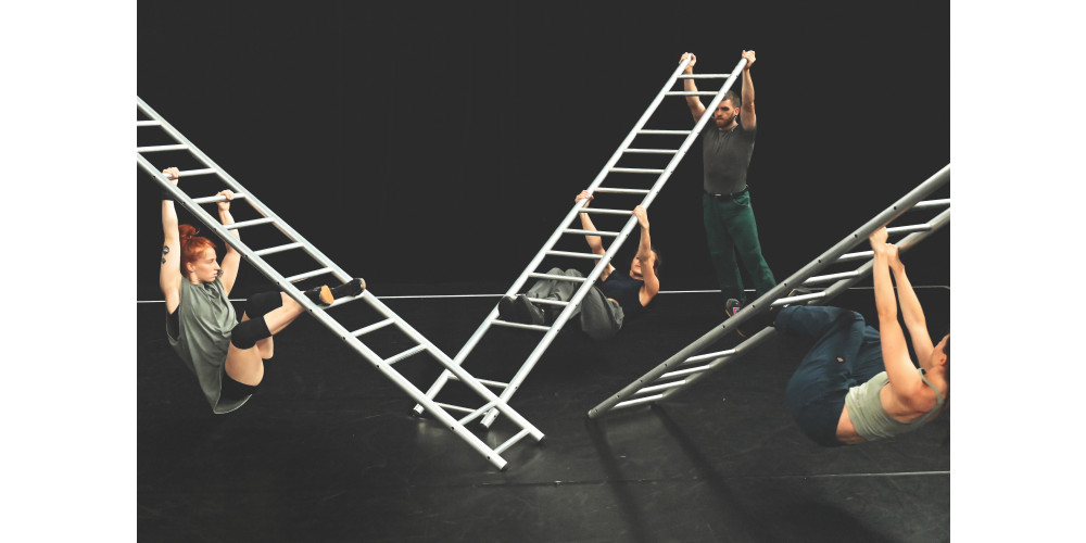 rei Performer*innen hängen mit Händen und Füßen unter schrägen Leitern in einem schwarzen Bühnenraum. In der Bildmitte liegen die Enden der drei Leitern auf dem Boden. Die oberen Enden der vorderen Leitern verschwinden rechts und links im Bildausschnitt. Das Ende der hinteren Leiter wird von einem vierten Performer mit gestreckten Armen über den Kopf gehalten. Die Performer*innen sind in dunklen grün und grau Tönen gekleidet und eine von ihnen trägt Knieschoner.