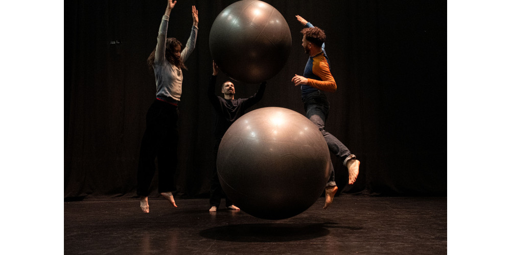 Foto: Drei Performer*innen interagieren im schwarzen Bühnenraum mit zwei großen grauen Bällen. Die vorderen beiden befinden sich im Sprung, alle drei in dynamischen Posen. Die beiden Bälle sind ebenfalls in der Luft, wirken durch die Perspektive unterschiedlich groß und übereinander. Die Performer*innen sind barfuß.