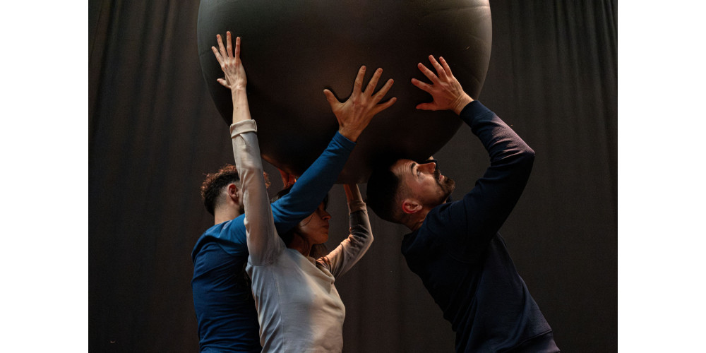 Foto: Drei Performer*innen stehen hintereinander und halten gemeinsam einen großen, grauen Ball über ihren Kopf. Nur die Oberkörper sind sichtbar. Die beiden linken Personen stehen eng und haben ihre Arme überkreuzt. Die Person rechts schaut nach oben. Sie tragen von links nach rechts blau, weiß und grau. Der Hintergrund ist schwarz.