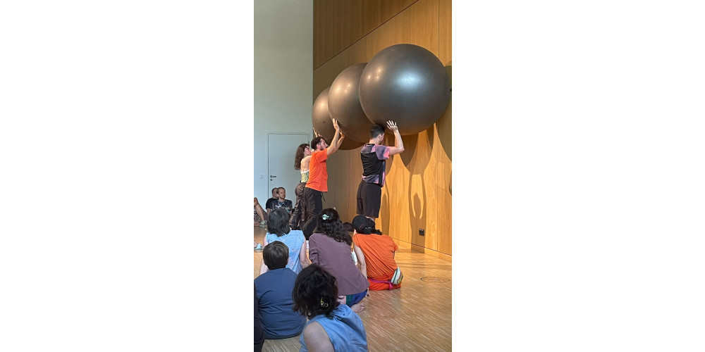 Foto: Drei Performer*innen halten große graue Bälle über ihren Köpfen rechts an die Wand. Auf dem Parkettboden sitzen acht Personen unterschiedlichen Alters und schauen zu. Die Performer*innen tragen schwarze Hosen und unterschiedliche Oberteile.