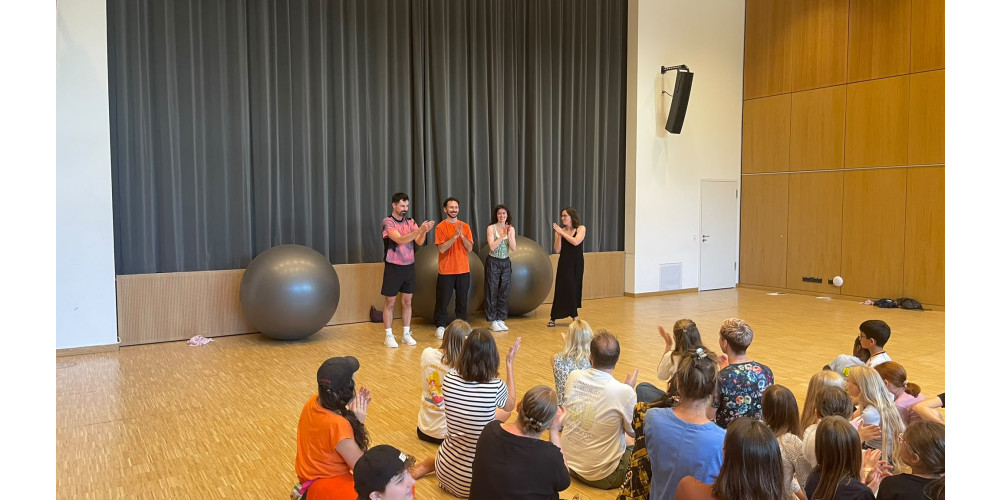 Foto: Schlussapplaus. Drei Perfomer*innen stehen vor einem Vorhang und klatschen. Daneben steht eine weitere Person im schwarzen Kleid und klatscht für sie. Das Publikum sitzt auf dem Fußboden und applaudiert. Die drei großen Bälle im Hintergrund gehen den Performer*innen bis zur Brust.