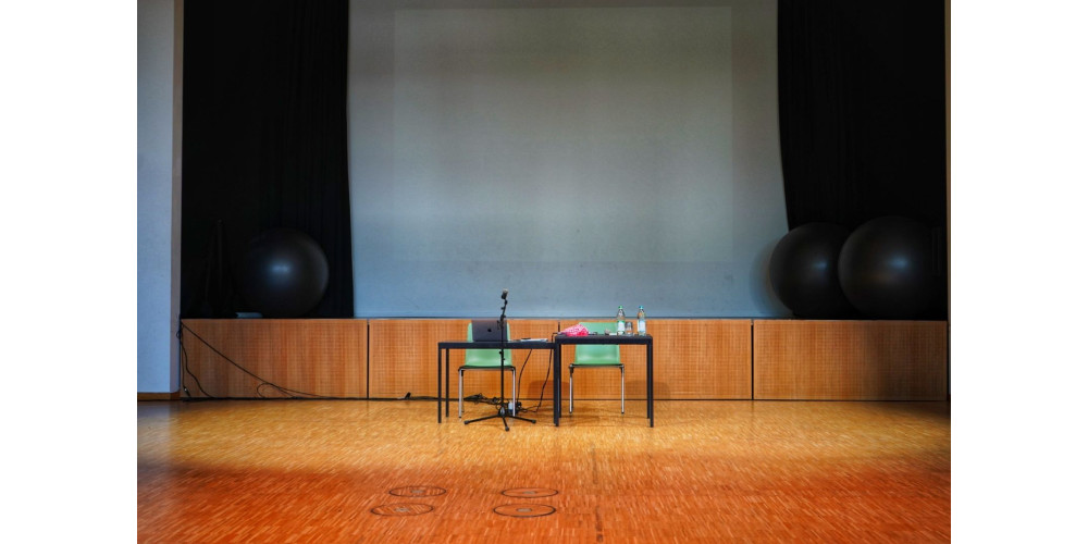 Foto: Blick auf die Bühne im Saal des Kulturzentrums Luise. Zwei schwarze Tische mit grünen Stühlen stehen auf dem Parkettboden vor einer weißen Leinwand von schwarzen Vorhängen gerahmt. Im Hintergrund liegen drei große graue Bälle. Auf den Tischen liegen Materialien und zwei Wasserflaschen. Davor steht ein Mikrofonständer.