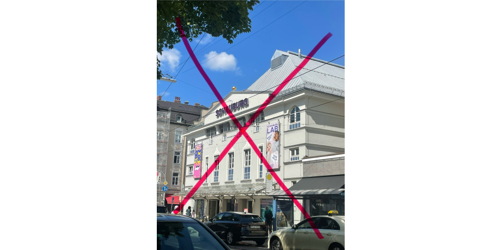 Die Frontansicht eines hellen Theatergebäudes mit der Aufschrift „Schauburg“ mit einem roten X übermalt. Davor stehen Autos. Im Hintergrund ist der blaue Himmel zu sehen.