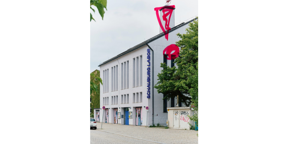 Foto: Die Frontansicht eines weißen Gebäudes mit der lila Aufschrift „Schauburg Labor“ mit einem rosa Ausrufungszeichen übermalt. Auf der rechten Seite verdecken Bäume schwarze vertikale Linien an der Gebäudefront. Der Himmel ist wolkenverhangen.