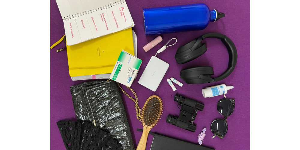 Foto: Auf lila Untergrund liegen folgende Gegenstände verstreut: eine blaue, metallene Wasserflasche, große, schwarze Kopfhörer, Augentropfen, eine Sonnenbrille, ein lila Hustenbonbon, ein schwarzes Fernglas, zwei Tampons, eine Haarbürste, eine weiße Powerbank, ein rosa Lippenbalsam, eine Packung Ibuprofen, eine schwarze Handtasche, ein aufgeklappter schwarzer Fächer, Notizbücher in gelb und schwarz.