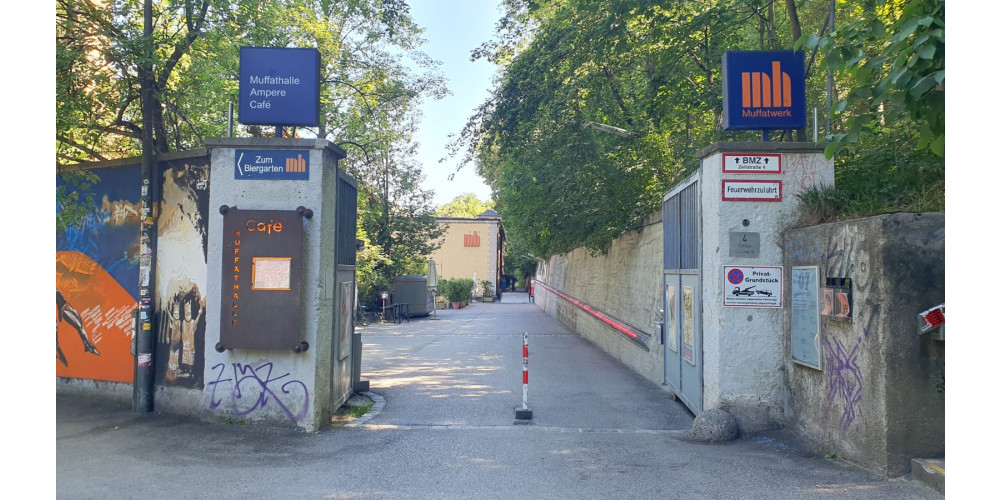 Foto: Eine Mauer mit einem offenen Tor mit einem Poller in der Mitte. Durch das Tor erstreckt sich ein leerer Platz an dessen Ende das Muffatwerk steht, ein gelbes Gebäude mit einem orangenen Logo an der Fassade. Der Himmel ist blau und viele Bäume werfen Schatten auf den Weg. Außen an der Mauer sind viele Schilder angebracht, die auf das Muffatwerk, sowie einen Biergarten, ein Café, eine Feuerwehrzufahrt und ein Privatgrundstück hinweisen.