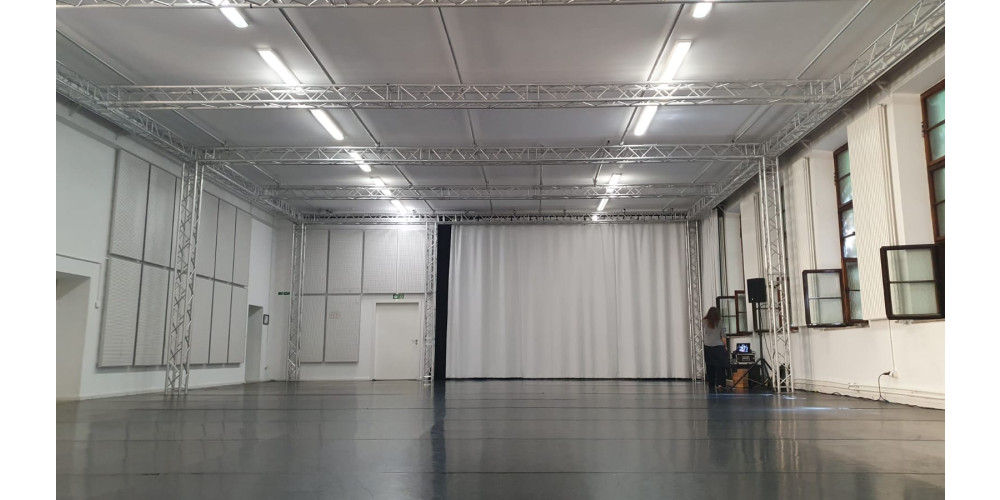 Foto: Ein großes, leeres Tanzstudio mit schwarzem Tanzteppich und weißen Wänden und Decken. Die Lichtröhren an der Decke sind eingeschaltet und werden gekreuzt von technischem Gerüst. Die Fenster auf der rechten Seite sind geöffnet. In der Ecke steht eine Person mit mittellangen braunen Haaren mit dem Rücken zur Kamera.